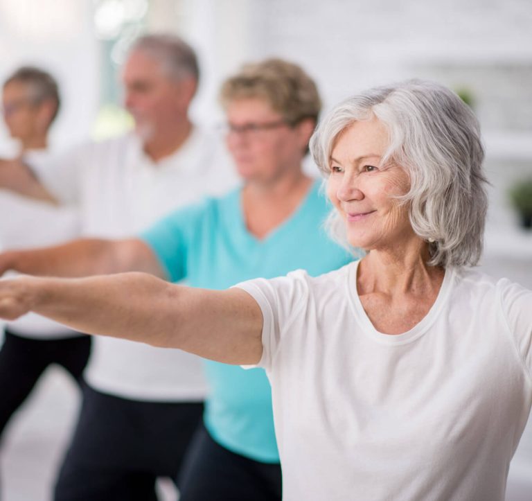 A multi-ethnic group of adult men and women are indoors in a fitness studio. They are wearing casual clothing while at a yoga class. A senior Caucasian woman is smiling while stretching out her arms.