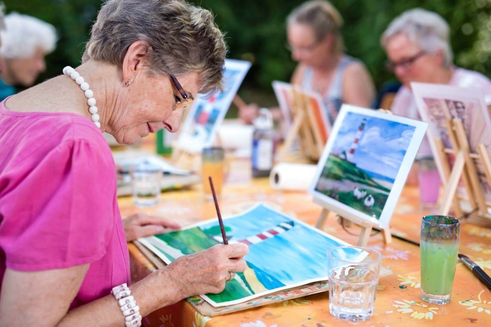 Art therapy for senior ladies, group of women painting the picture of lighthouse from the watercolor template  sitting at one table outdoors in the park.